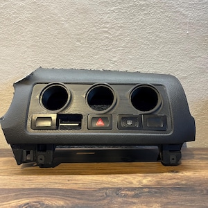 Puede incluir: Un componente negro del salpicadero de un coche con tres aberturas circulares para portavasos y varias ranuras rectangulares para botones. Es visible un botón de peligro triangular rojo. El componente es de plástico texturizado.