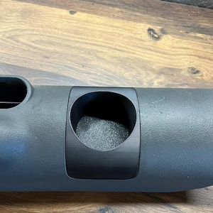Op de afbeelding: Een zwarte plastic bekerhouder met een grijs, getextureerd oppervlak. De bekerhouder heeft een ronde opening en is ontworpen om een beker van standaardformaat te passen.