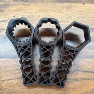 May include: Three black 3D-printed wrenches with different head shapes: a gear, a rounded shape, and a hexagon. The handles have a lattice design. The tools are arranged on a wooden surface.