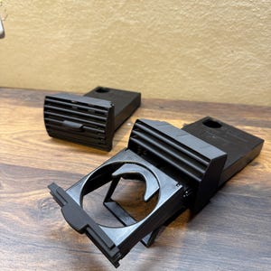 Puede incluir: Conjunto negro de portavasos y ventilación de coche. El portavasos está abierto. La ventilación tiene lamas horizontales. Los artículos están sobre una superficie de madera.