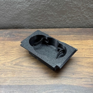 Peut inclure: Un porte-gobelet de console de voiture noir et rectangulaire avec deux compartiments circulaires. Le porte-gobelet est fait d'un matériau texturé et repose sur une surface en bois.