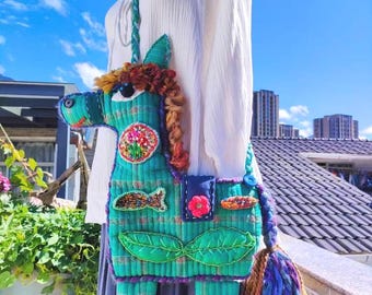 Può includere: Borsa a tracolla fatta a mano a forma di cavallo. La borsa è principalmente turchese con dettagli ricamati colorati, tra cui un fiore, foglie e un pesce. Ha una tracolla intrecciata e un nappa, perfetta per trasportare l'essenziale.