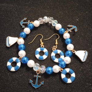May include: A nautical-themed jewelry set featuring a bracelet and earrings. The bracelet is made of blue, white, and clear beads with blue anchor and sailboat charms. The earrings have gold hooks and blue and white life preserver charms.