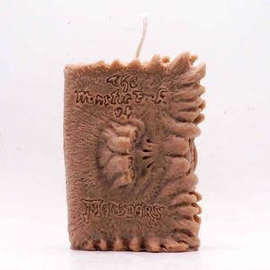 May include: Brown candle shaped like a book with the text "The Monster's of Monsters" embossed on the front. The candle has a textured, aged appearance and a white wick.