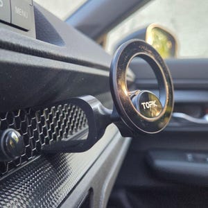 May include: A black circular magnetic phone mount with the brand name TOPK printed in white on the center. The mount features a U-shaped arm that attaches to the horizontal honeycomb-patterned air vent of a vehicle dashboard interior.