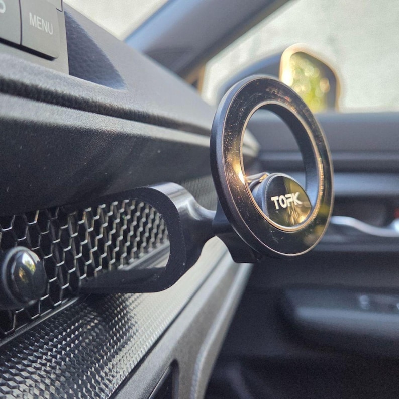 May include: A black circular magnetic phone mount with the brand name TOPK printed in white on the center. The mount features a U-shaped arm that attaches to the horizontal honeycomb-patterned air vent of a vehicle dashboard interior.
