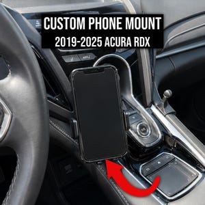 Può includere: Un supporto per telefono nero installato nell'abitacolo di un veicolo, con uno smartphone inserito. Il testo sull'immagine recita CUSTOM PHONE MOUNT 2019-2025 ACURA RDX. Una freccia rossa indica la base del supporto, evidenziandone la posizione.