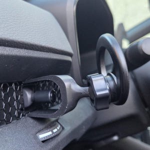 May include: A matte black car phone mount attached to a vehicle dashboard vent. The mount features a U-shaped bracket secured to the honeycomb-patterned vent grille, connected to a circular ring holder by a central adjustable knob mechanism.