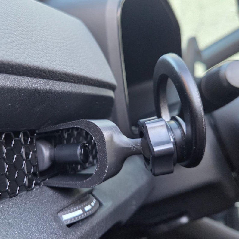 May include: A matte black car phone mount attached to a vehicle dashboard vent. The mount features a U-shaped bracket secured to the honeycomb-patterned vent grille, connected to a circular ring holder by a central adjustable knob mechanism.