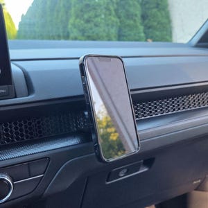 May include: A black smartphone mounted vertically on a car dashboard. The device features a slim black frame and a reflective screen surface. It is positioned directly in front of a dark gray dashboard panel with a distinct hexagonal honeycomb pattern.