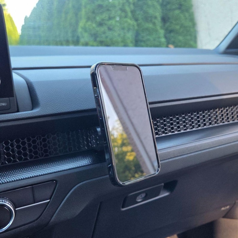 May include: A black smartphone mounted vertically on a car dashboard. The device features a slim black frame and a reflective screen surface. It is positioned directly in front of a dark gray dashboard panel with a distinct hexagonal honeycomb pattern.