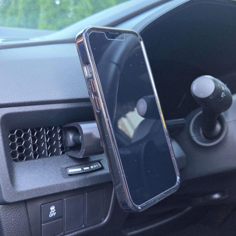 May include: A black smartphone in a clear protective case mounted to a car air vent. The mount is attached to a horizontal vent with a hexagonal grid pattern. The phone is positioned vertically, displaying a dark screen with a visible notch at the top.
