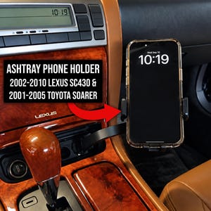 May include: A black phone holder mounted in the ashtray area of a vehicle interior with wood grain trim. The holder supports a smartphone displaying the time 10:19. Text on the image reads: ASHTRAY PHONE HOLDER 2002-2010 LEXUS SC430 & 2001-2005 TOYOTA SOARER.