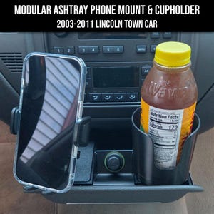 May include: A black modular ashtray phone mount and cupholder for a 2003-2011 Lincoln Town Car. The device is installed in the vehicle console, featuring a side-mounted smartphone holder and a circular cupholder containing a plastic beverage bottle.