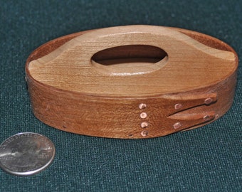 Könnte beinhalten: Eine kleine, ovale Holzkiste mit einem Griff oben. Die Kiste ist aus hellem braunem Holz und hat eine dunkelbraune Beize. Es gibt kleine Kupfernägel am Rand der Kiste.