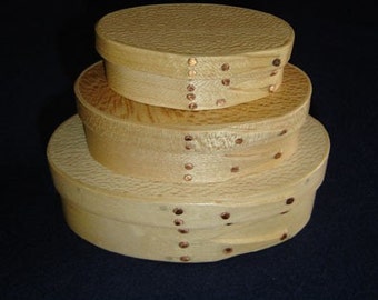 Könnte beinhalten: Drei ovale Holzkisten, ineinander verschachtelt, mit Kupfernägeln an den Rändern. Die Kisten sind aus hellem Holz und haben eine natürliche Oberfläche.