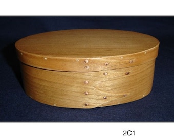 Könnte beinhalten: Eine ovale Holzkiste mit hellbrauner Oberfläche und Kupfernägeln am Rand. Die Kiste ist aus Holz und hat eine glatte Oberfläche.