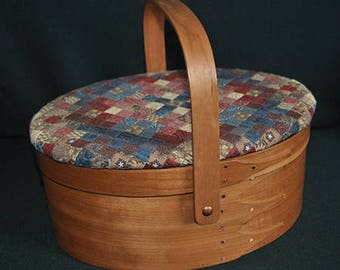 Könnte beinhalten: Eine ovale Nähbox aus Holz mit einem Scharnierdeckel und einem Stoffbezug. Die Box hat einen Holzgriff und ist aus hellbraunem Holz gefertigt.