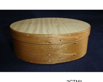 Könnte beinhalten: Eine runde Holzkiste mit einem hellen Holzdeckel und einem dunkleren Holzboden. Die Kiste hat ein schlichtes Design und ist mit kleinen Kupfernägeln verziert.