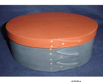 Könnte beinhalten: Eine dunkelblaue ovale Holzkiste mit einem roten Deckel und Messingnägeln. Der Deckel hat ein dekoratives Design mit zwei Lederriemen.