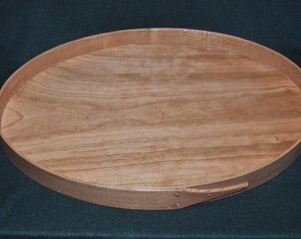 Könnte beinhalten: Ein rundes Holztablett mit einer natürlichen Holzoptik. Das Tablett hat einen erhöhten Rand und ist aus Kirschholz gefertigt.