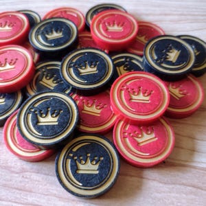 May include: A pile of red and black game pieces with a gold crown design. The pieces are round and have a raised edge.