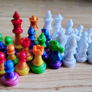 Könnte beinhalten: Ein farbenfrohes Schachspiel mit Figuren in verschiedenen Farben, darunter Rot, Orange, Grün, Blau und Lila. Das Set enthält Könige, Damen, Türme, Läufer, Springer und Bauern. Die Figuren sind auf einer hellbraunen Holzoberfläche angeordnet.