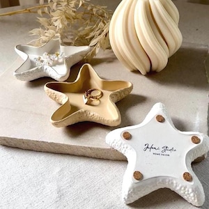 Puede incluir: Tres platos de cerámica en forma de estrella de mar en blanco y beige. Los platos tienen forma de estrella de mar y una superficie texturizada. El plato más grande tiene el texto "Jufeni Studio Home Decor" en la parte inferior.