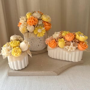 May include: Three floral arrangements in white containers. The flowers are in shades of yellow, orange, and white, resembling roses, daisies, and other blooms. The arrangements sit on a neutral-toned surface, creating a soft, inviting aesthetic.