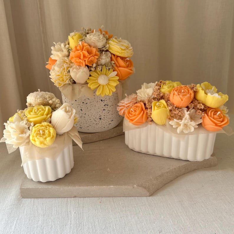 May include: Three floral arrangements in white containers. The flowers are in shades of yellow, orange, and white, resembling roses, daisies, and other blooms. The arrangements sit on a neutral-toned surface, creating a soft, inviting aesthetic.