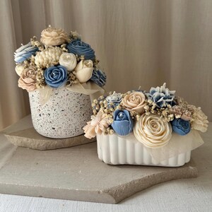 May include: Two floral arrangements in white containers. One is a speckled, cylindrical vase, and the other is a rectangular, ribbed container. Both feature pastel-colored, sculpted flowers in shades of blue, cream, and peach.