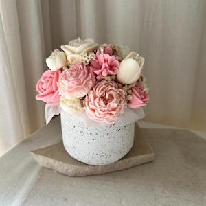 May include: A floral arrangement of flower-shaped candles in a speckled white ceramic pot. The candles are various shades of pink and white, resembling roses, peonies, and tulips. The pot sits on a small, rectangular stone.