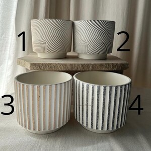 May include: Four ceramic planters in neutral tones. Two have a textured, swirling design, and two feature vertical stripes. The planters are cylindrical with a slightly flared base, suitable for indoor or outdoor use. The planters are ideal for succulents or small plants.