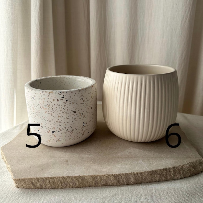 May include: Two ceramic planters. One is a small, speckled, off-white pot with pink and brown flecks. The other is a larger, cream-colored pot with vertical ribbed detailing. Both are on a light-colored stone surface.