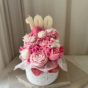 May include: A floral arrangement in a speckled white pot, featuring pink and white flowers, with three cream-colored candles spelling out "100". The flowers are arranged with pink accents and a white ribbon.