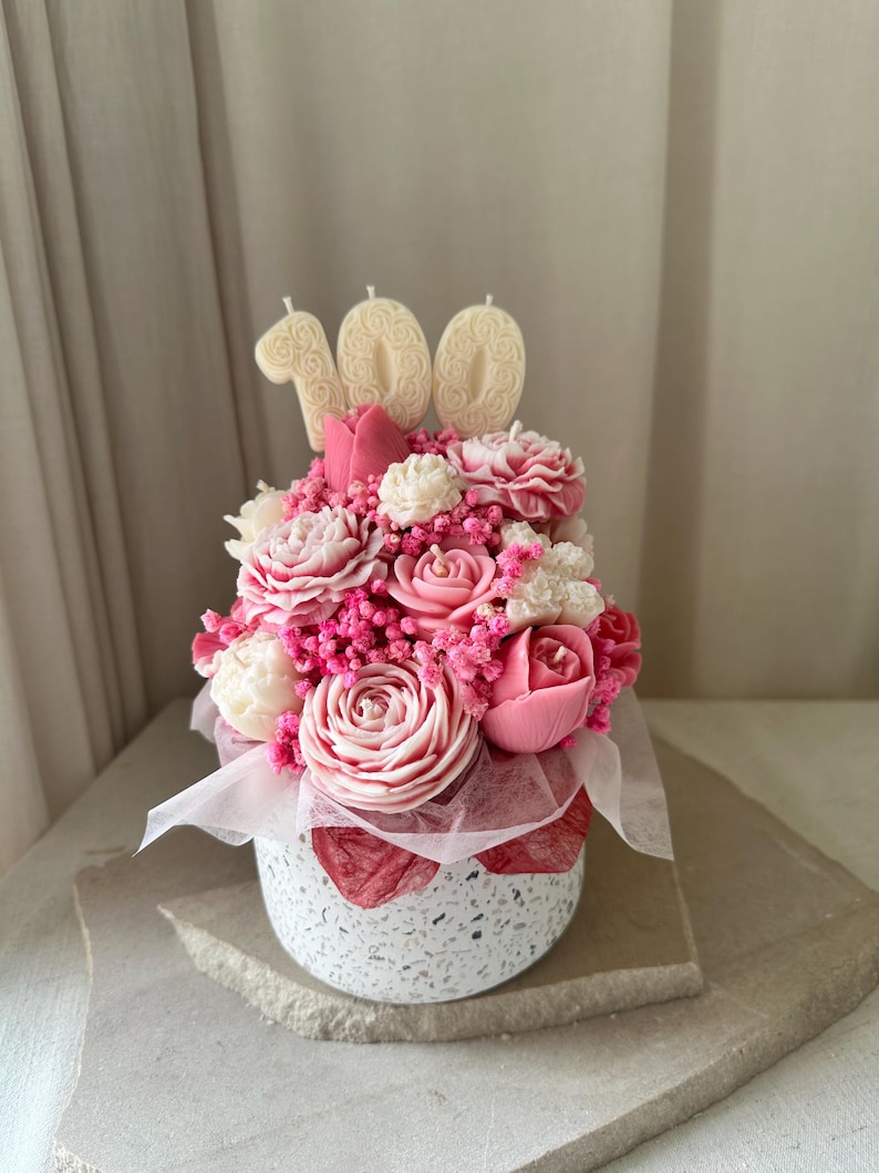 May include: A floral arrangement in a speckled white pot, featuring pink and white flowers, with three cream-colored candles spelling out "100". The flowers are arranged with pink accents and a white ribbon.