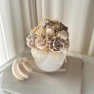 May include: A floral arrangement of carved soap flowers in shades of pink, cream, and brown, presented in a speckled white container. A smaller, swirled soap sculpture sits nearby. The soaps are arranged to resemble a bouquet.