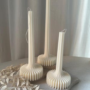 Peut inclure: Trois bougies blanches en forme de pilier avec un design unique et cannelé. Les bougies ont une base circulaire large avec un bord festonné.