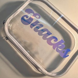 May include: Clear rectangular zippered pouch with the word "Snacks" in a gradient purple and blue font. The pouch has a white trim and zipper, and is made of a transparent material.