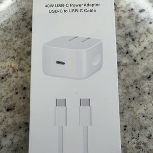 Puede incluir: Una caja de producto rectangular blanca con la etiqueta 40W USB-C Power Adapter y USB-C to USB-C Cable. El empaque presenta una ilustración gráfica de un adaptador de corriente cuadrado blanco con un puerto y un cable USB-C blanco.
