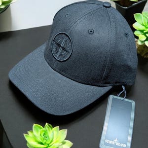 May include: A dark navy baseball cap with a curved peak. The cap has a circular emblem with a compass design. A tag with the text "STONE ISLAND" is attached. The cap is on a black surface with green plants.
