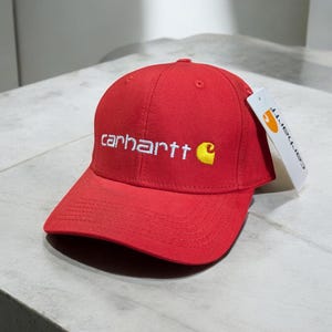 May include: A red baseball cap featuring the white embroidered word "carhartt" and a yellow "C" logo. A white tag with the brand name is attached. The cap is made of a textured fabric and rests on a light grey surface.