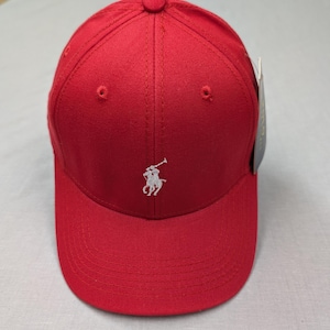 May include: A red baseball cap with a curved peak. The cap has a white embroidered polo player logo on the front. It features a button on the crown and a tag attached to the side.