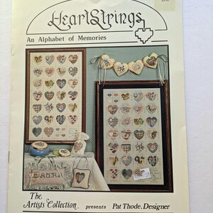 May include: A book cover titled "HeartStrings, An Alphabet of Memories" featuring framed cross-stitch heart designs. The cover includes the text "The Artists Collection presents Pat Thode, Designer". The image also shows a heart-shaped garland.