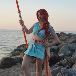 May include: A person in a cosplay outfit stands on a beach, holding an orange staff with white stripes. The outfit includes a light blue dress, a peach-colored sash, and red hair. The background shows the ocean and rocks.