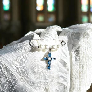 May include: A white pearl and silver safety pin with a blue crystal cross pendant. The pin is on a white lace fabric.
