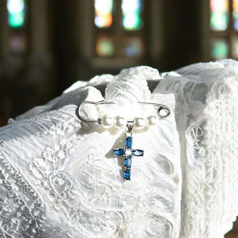 May include: A white pearl and silver safety pin with a blue crystal cross pendant. The pin is on a white lace fabric.