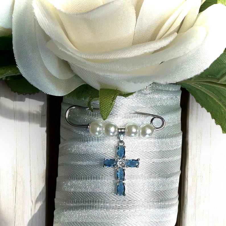May include: A white satin ribbon with a safety pin and a blue crystal cross charm. The charm is attached to a string of white pearls.