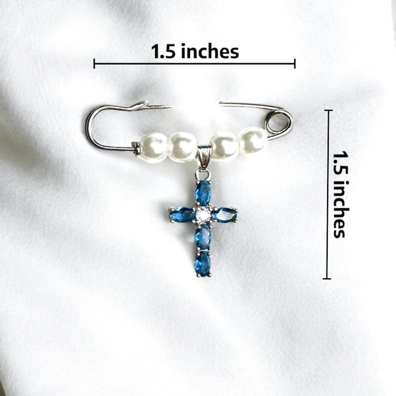 May include: A silver safety pin with a string of white pearls and a blue crystal cross pendant. The pin measures 1.5 inches in length and width.