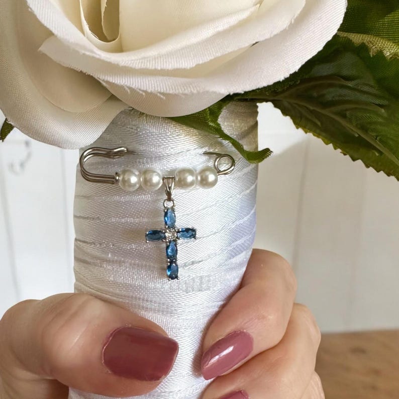 May include: A white bridal bouquet with a silver safety pin holding a blue crystal cross charm with white pearls.